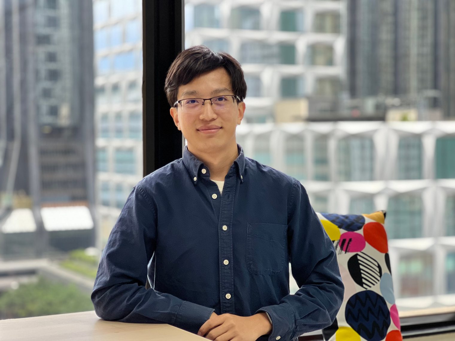 The author, half-length, in glasses and a dark blue shirt, smiling and leaning on a table by a window, with tall buildings outside