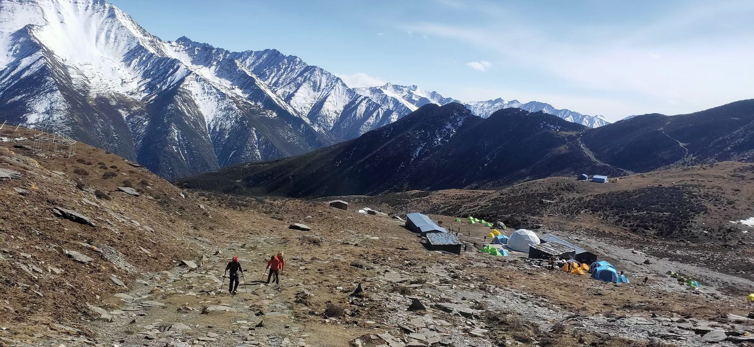 Tents and a few tin-roofed huts scattered across a valley below snow mountains; two climbers in red walk up a rocky path with trekking poles, and a line of snowy peaks stretches away behind