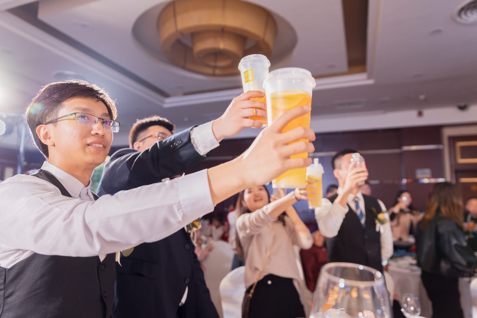 At a wedding banquet, a few men in suits and waistcoats and a woman raise cups of orange drink high in a toast