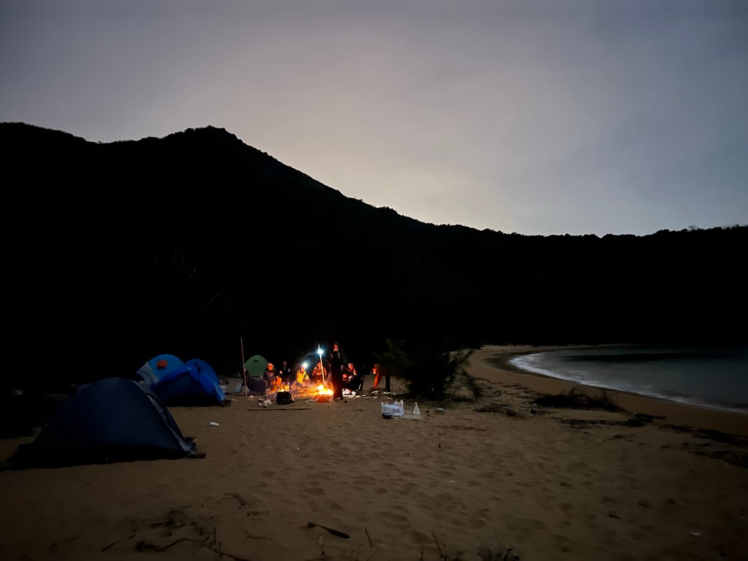 A beach at night: a group of people gathered round a campfire beside a few tents, a black hill behind them and a curving bay to the right