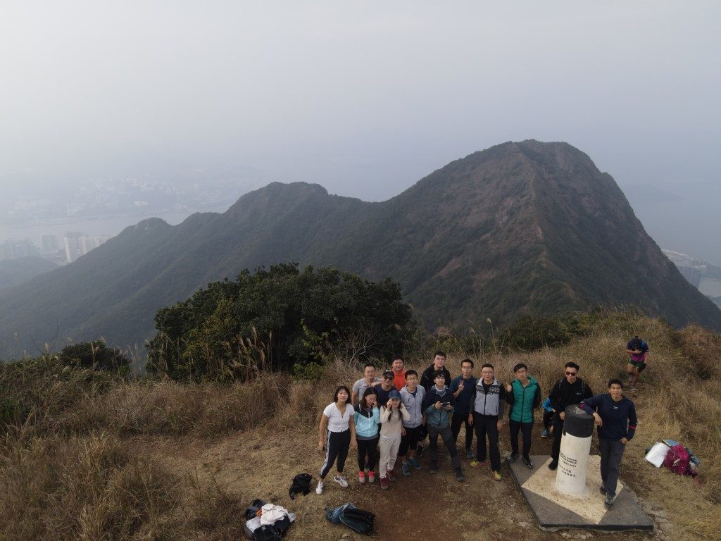 A group photo on a summit taken by drone: a dozen or more people stand by a trig pillar, with a ridge behind them and, faint in the haze, a city and the sea