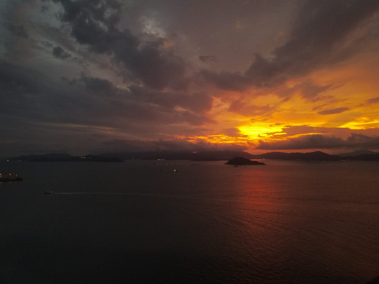 A sunset over the sea seen from the air: heavy clouds split by a band of red-gold light, a line of hills and a small island in the distance, and a few ship lights on the water