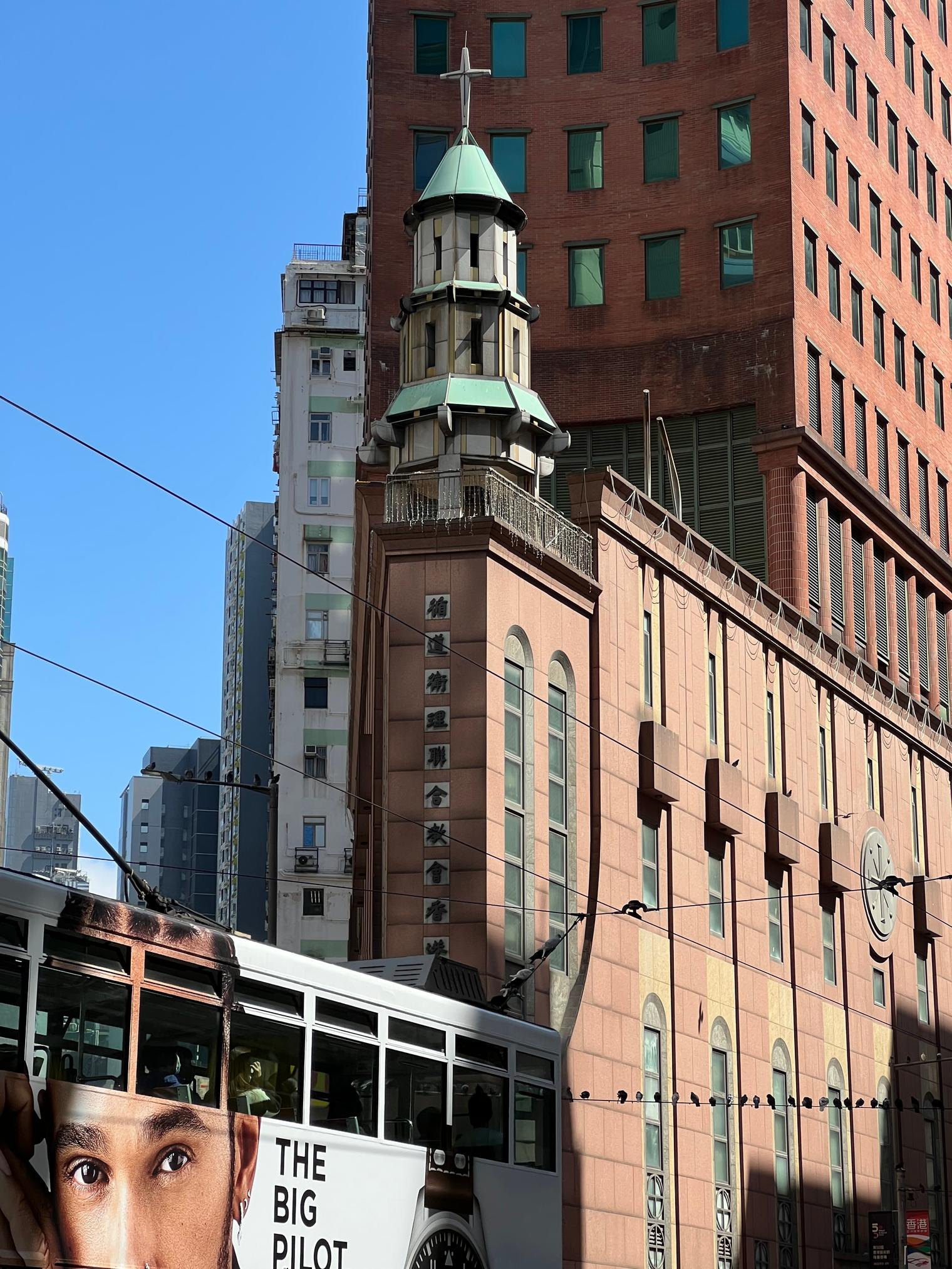 The green-roofed bell tower and red facade of a Methodist church, a tram passing in front and a row of pigeons on the overhead wires