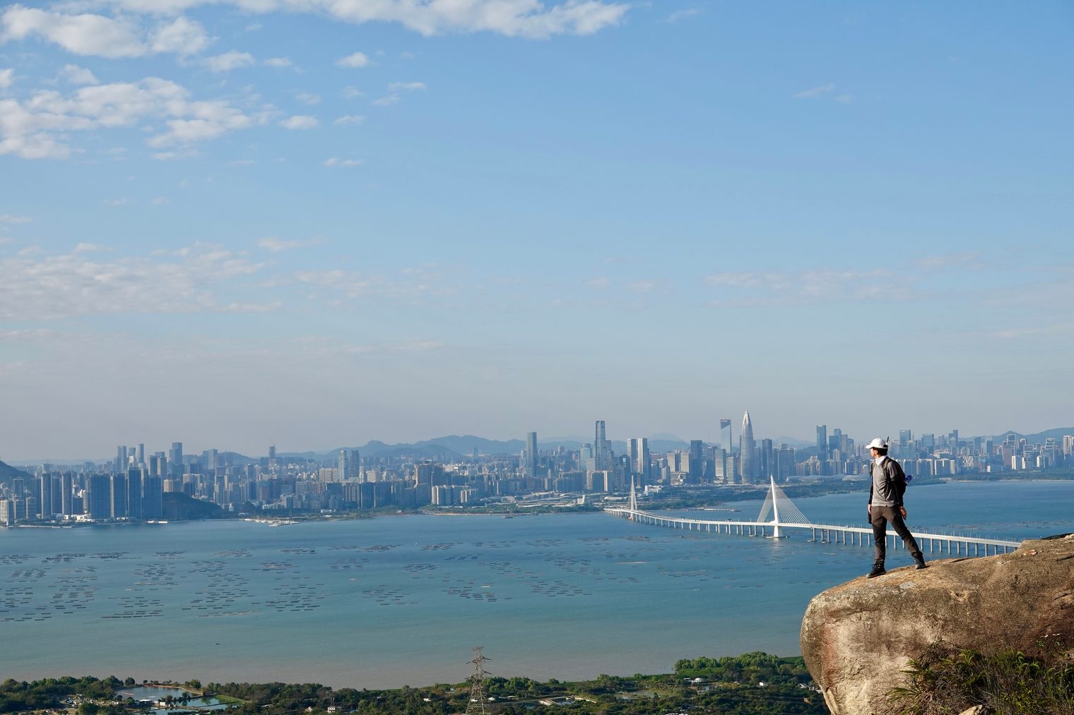 A hiker stands at the edge of a boulder, looking across the bay to the towers of Shenzhen and the Shenzhen Bay Bridge