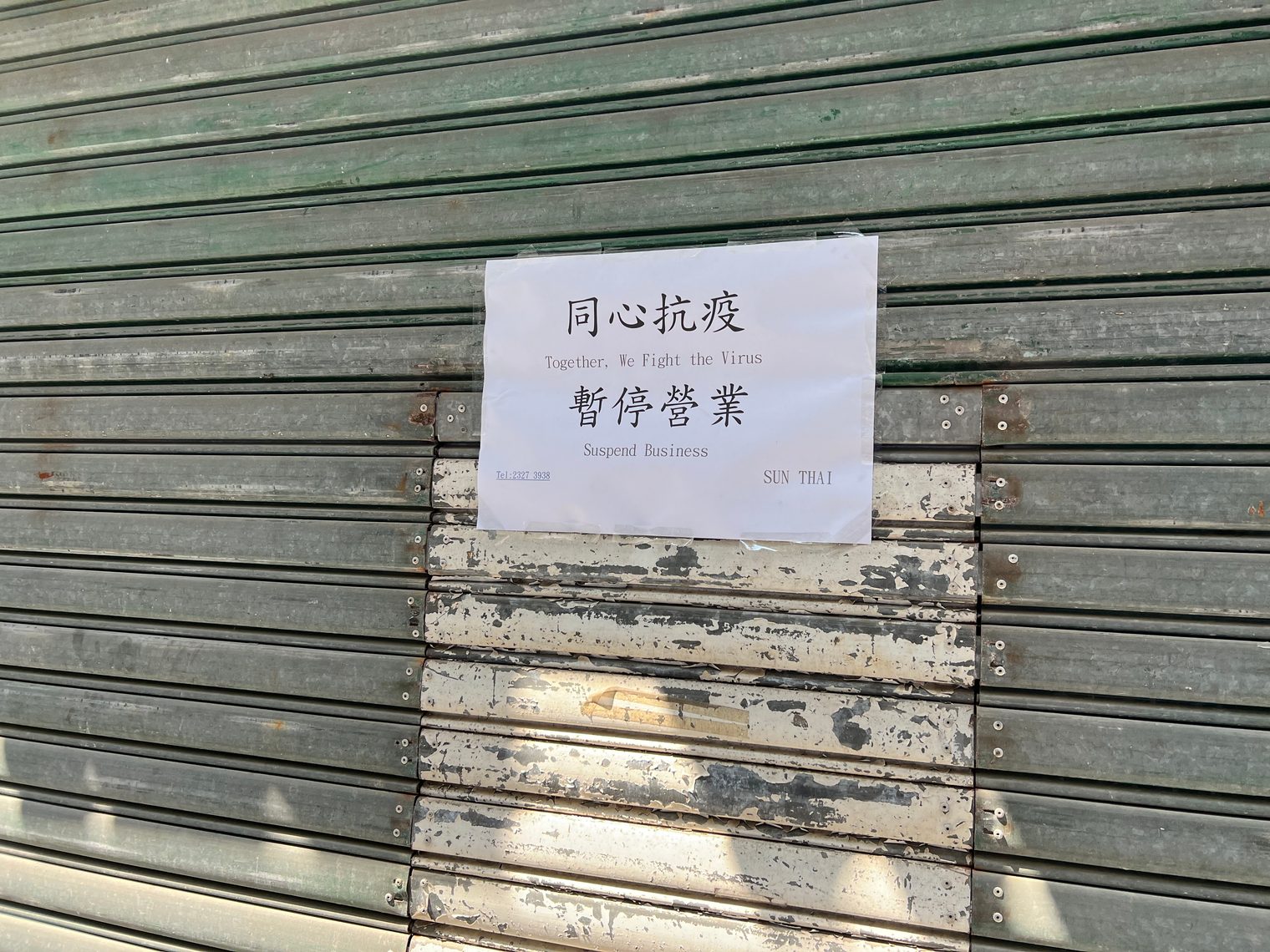 A notice taped to a closed green roller shutter: "Together, We Fight the Virus. Suspend Business"