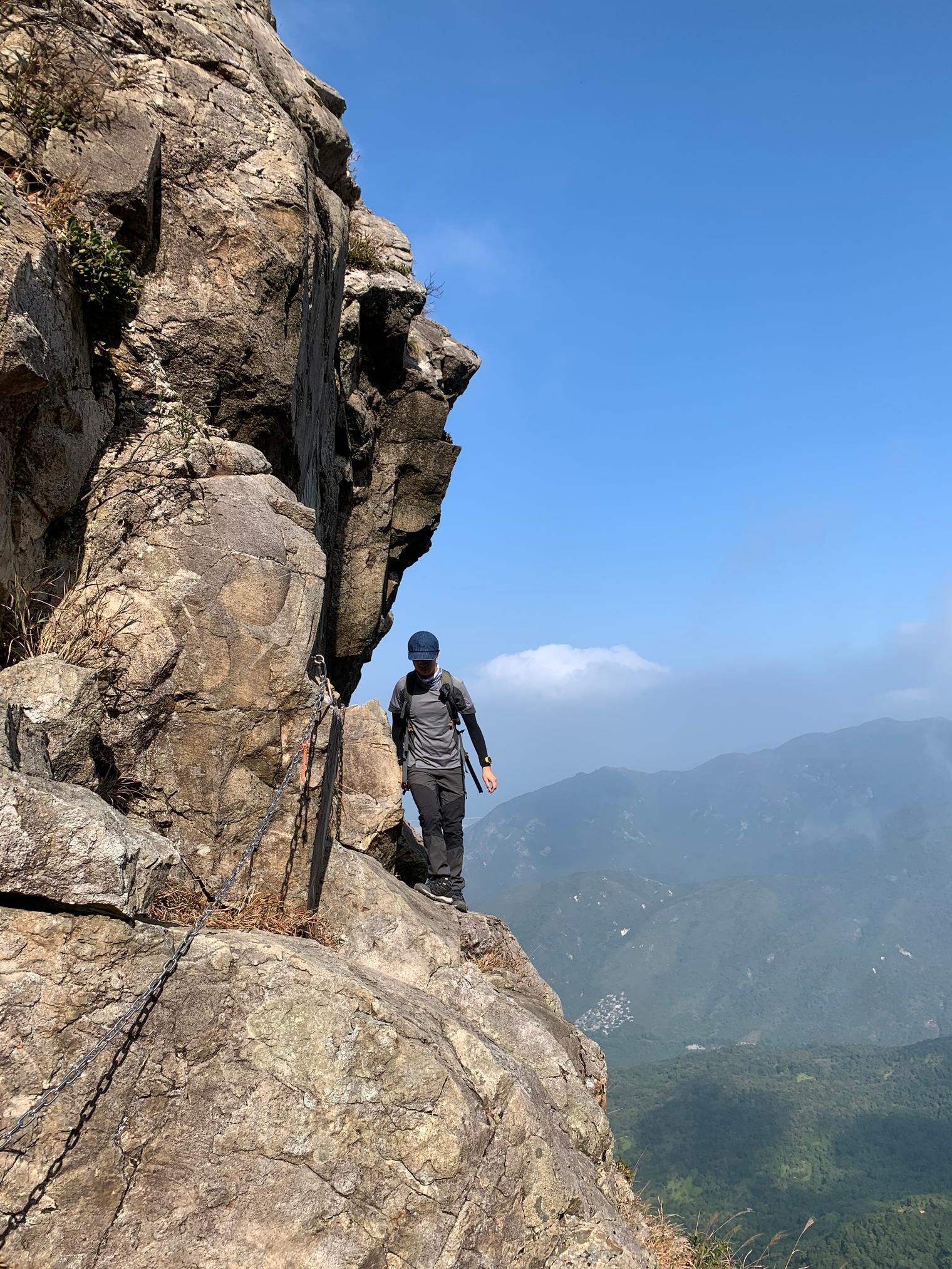 A hiker in a cap edges along a narrow rock ledge under a cliff, beside a chain, with hazy valleys and mountains beyond
