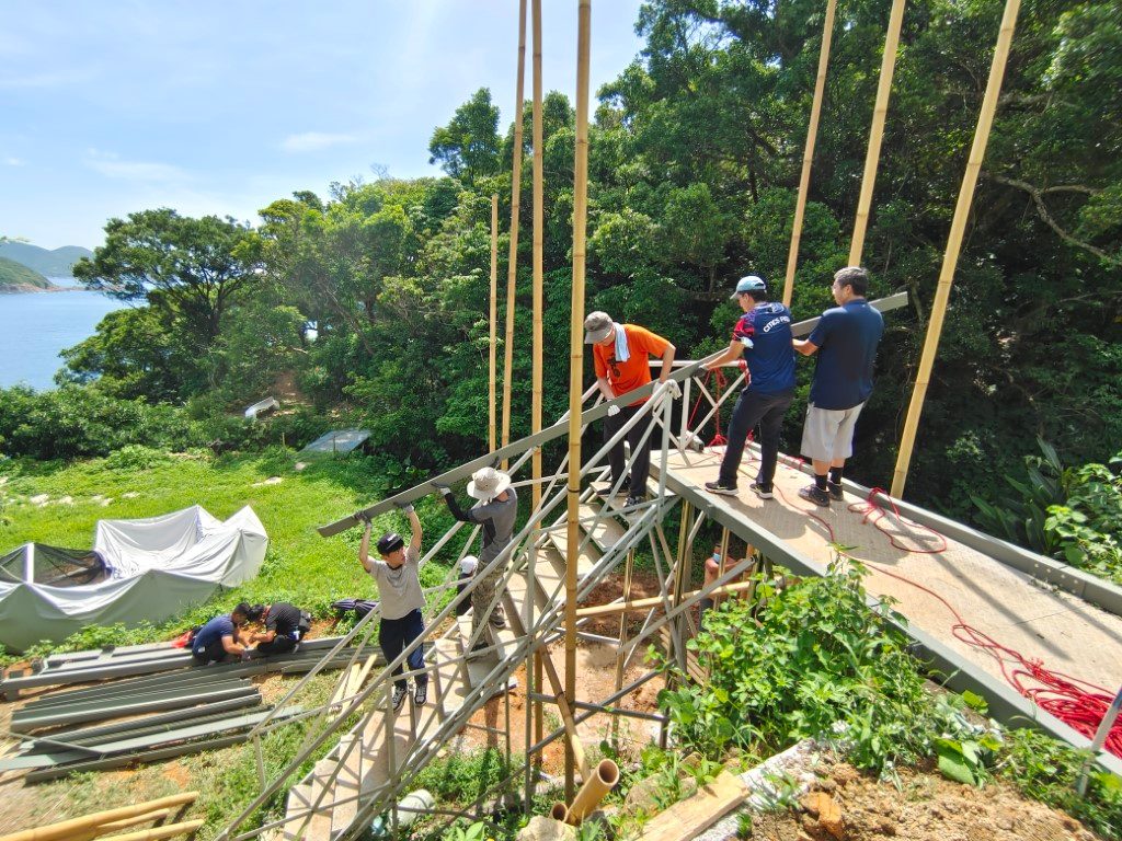 At a hillside farm by the sea, a group of people work together to raise a structure of steel frames and bamboo poles
