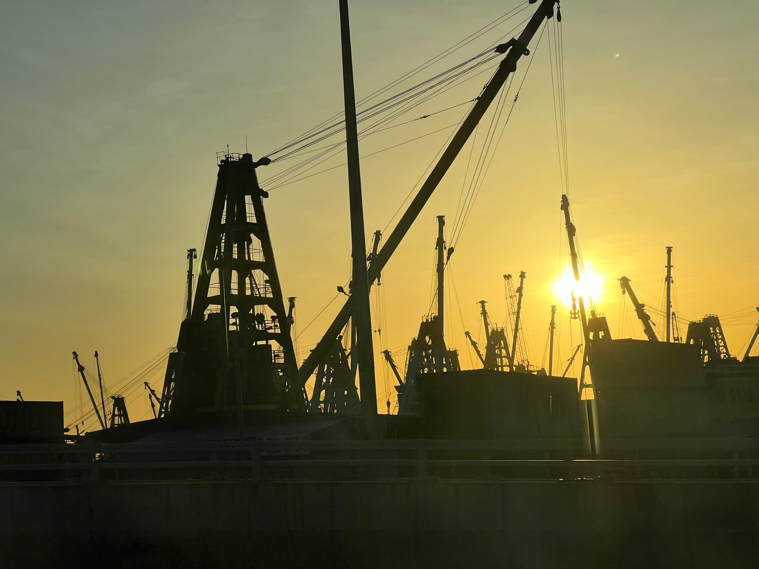 The booms and masts of crane barges in silhouette against the setting sun