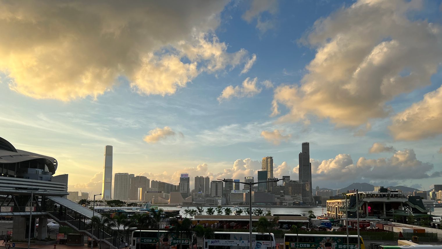 Victoria Harbour from outside Exhibition Centre station: evening clouds over the International Commerce Centre and the Kowloon skyline across the water, with a lamp post and buses in front