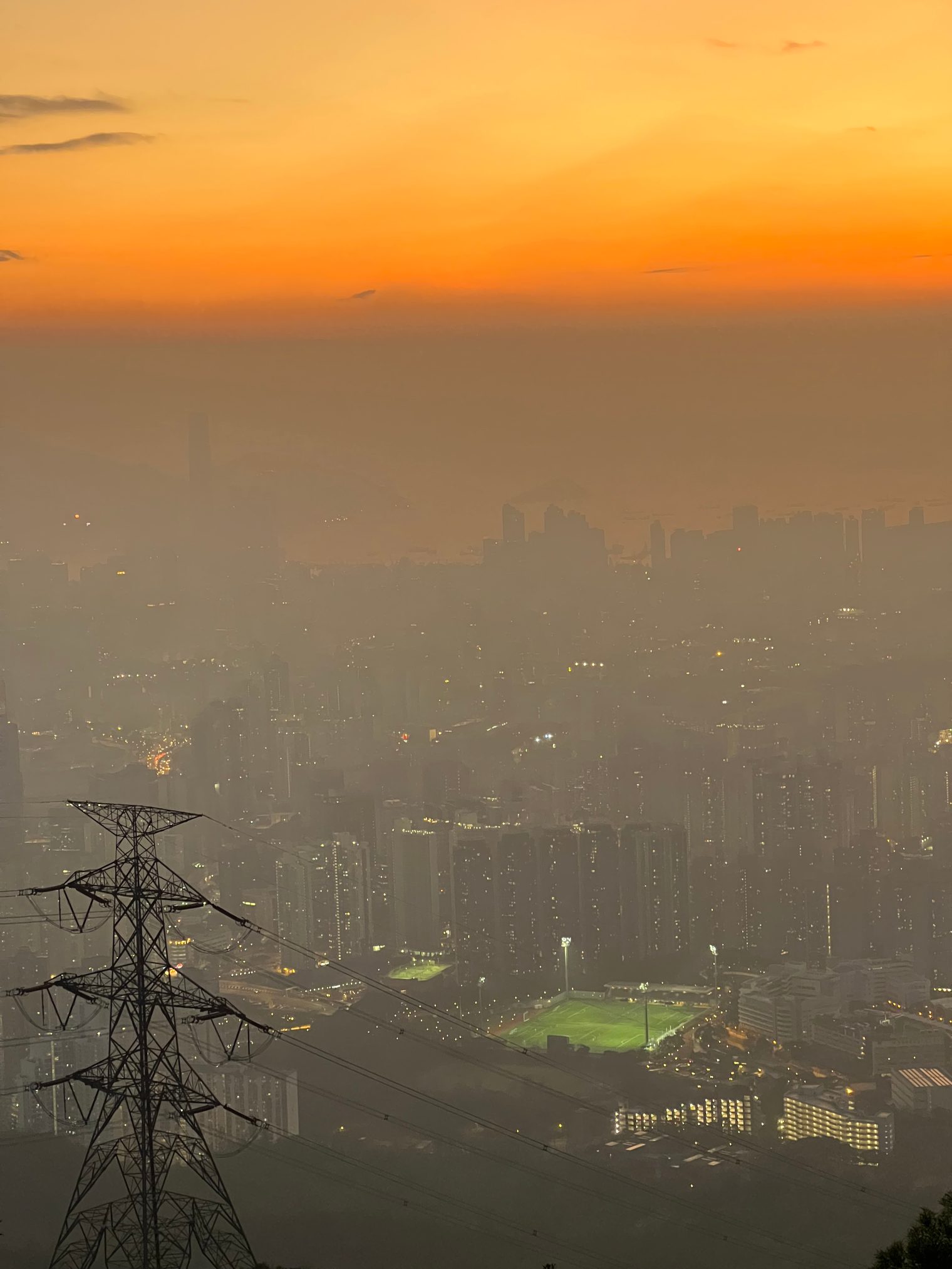 The city in haze under an orange sky, a floodlit football pitch below and a pylon in the lower left