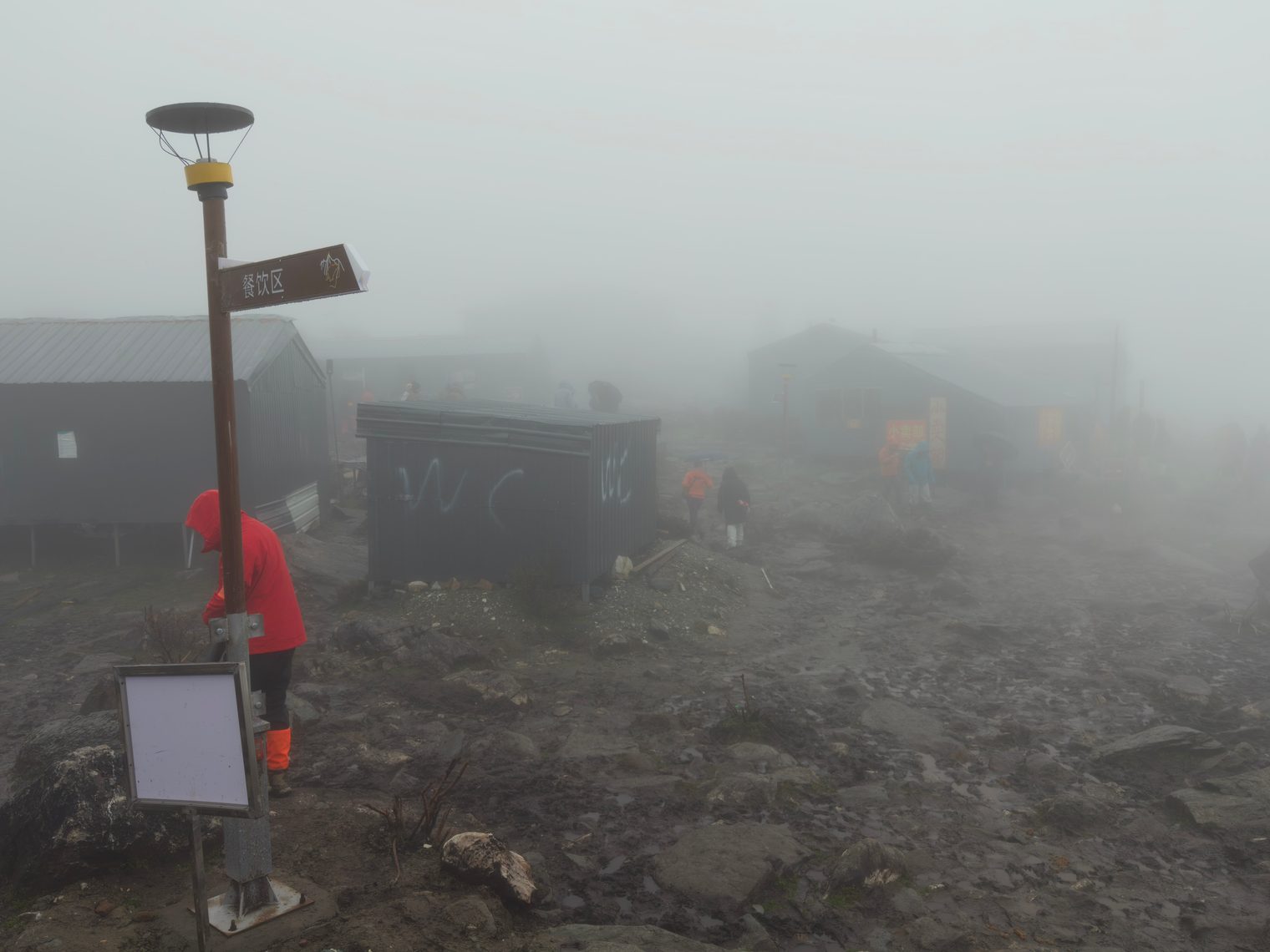 Base camp in fog: corrugated metal huts, a signpost, and a toilet block