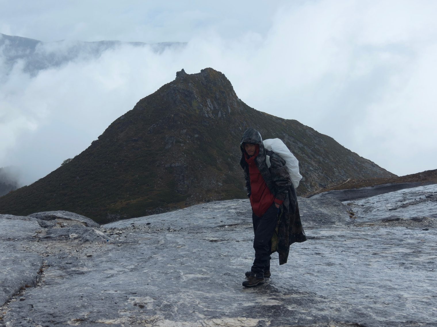 A porter carrying a white load sack across grey rock slabs below a peak