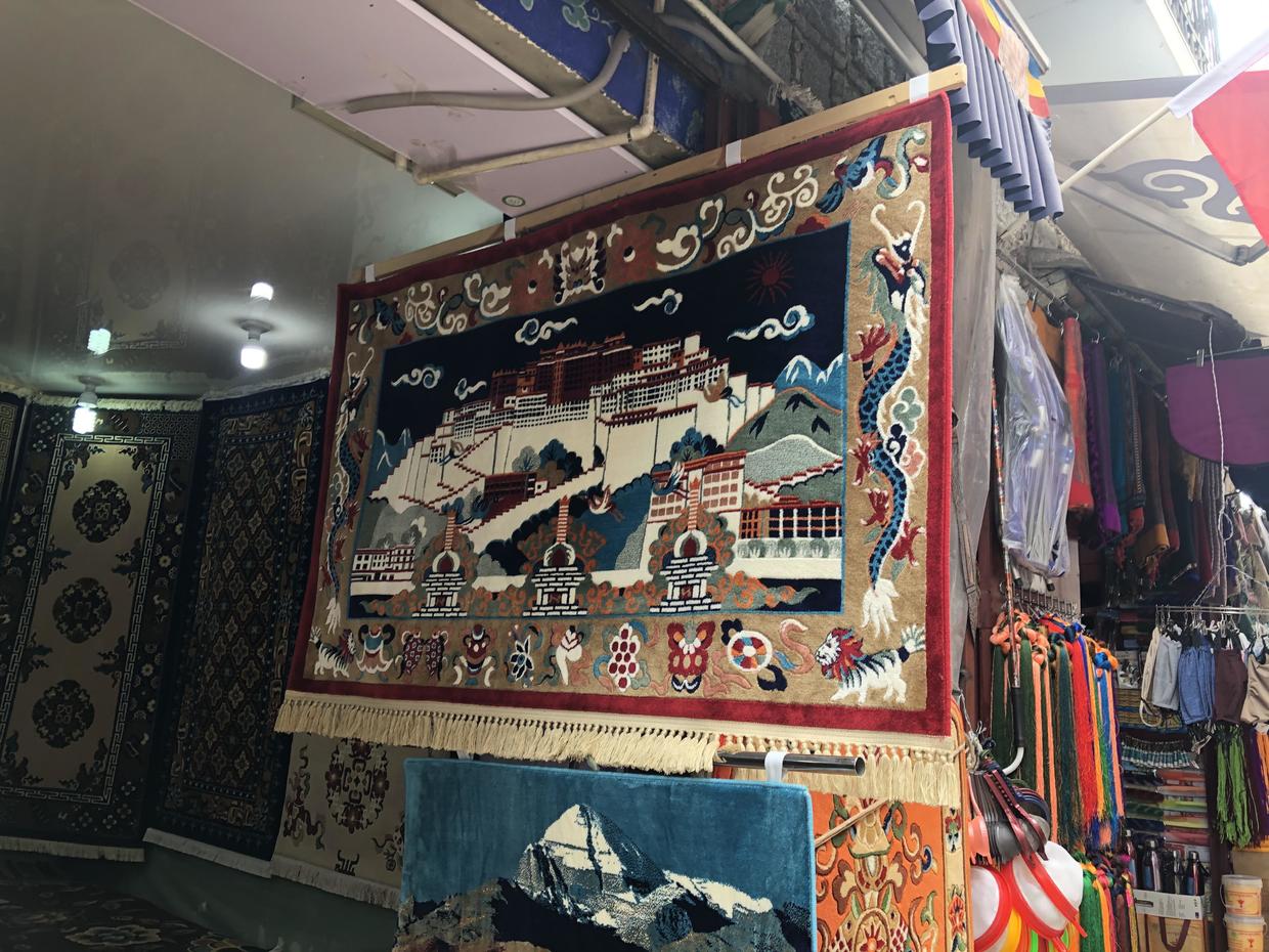 Tibetan rugs hanging in a shop: one woven with the Potala Palace, white stupas, dragons and a snow lion, another below it with a snow mountain