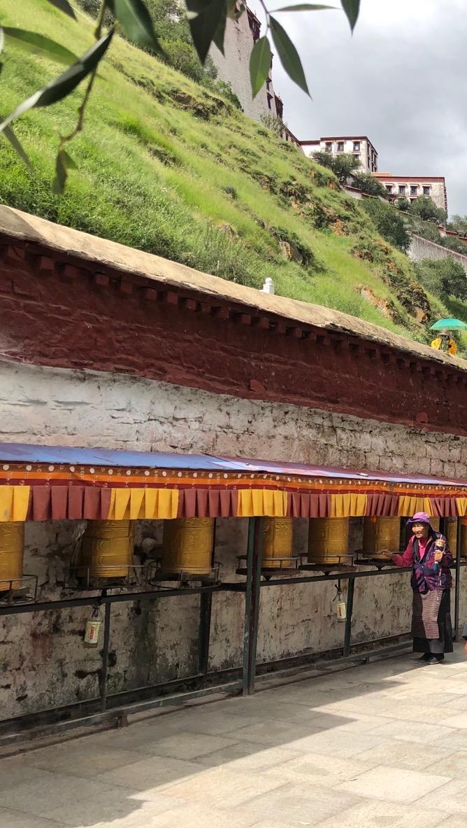 A row of golden prayer wheels along a red-and-white wall below a grassy slope with palace buildings above; an elderly woman in a hat is turning them