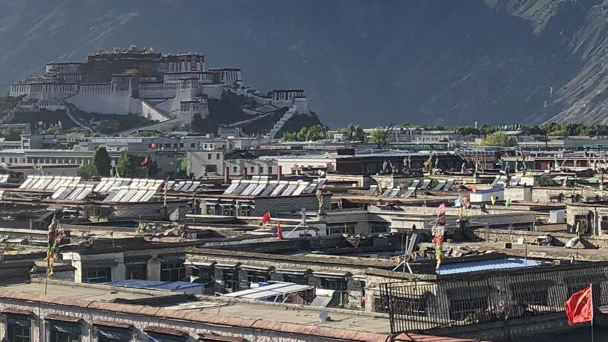 Lhasa seen from above: the Potala Palace on its hill in the distance, and in front, low flat-roofed buildings lined with solar panels and flying prayer flags and Chinese national flags
