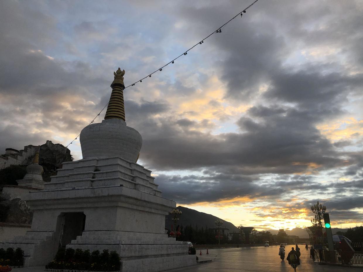 A white stupa by the road under evening clouds; a line hung with small bells runs up from its golden spire, another small white stupa stands on the hill to the left, and a few scooters ride along the road