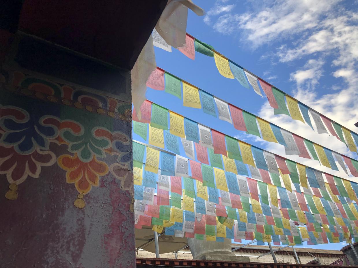 Rows of blue, white, red, green and yellow prayer flags strung out beside a painted pillar, under a blue sky with white clouds