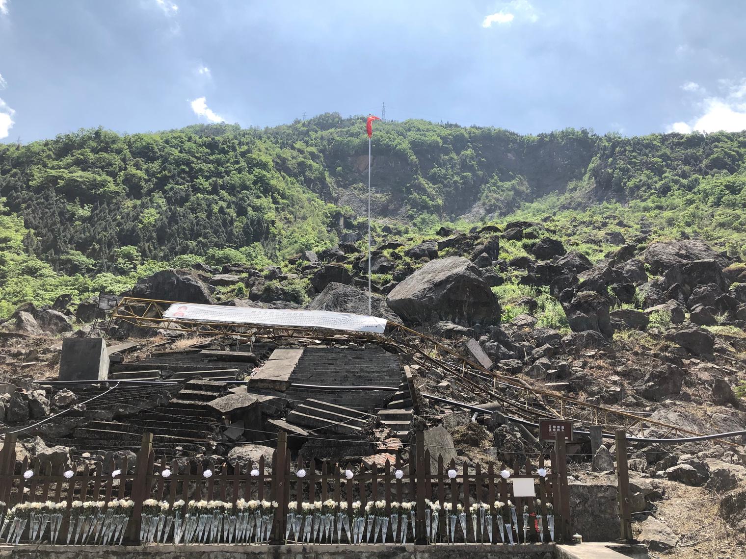 An earthquake ruin at the foot of a hillside, with white flowers laid before broken steps and the national flag standing in the middle