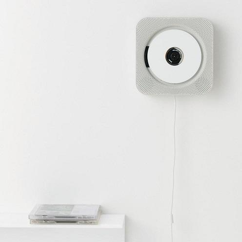 Original figure: a square white CD player on a white wall, with a perforated front, central disc, and a pull cord hanging below; CDs rest on a shelf at lower left